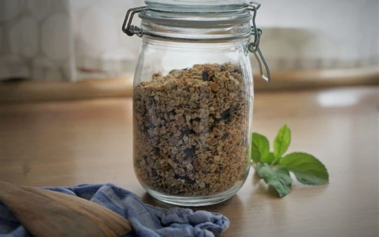 homemade granola in a glass container, a wooden spatula, a towel and some apple tree leaves