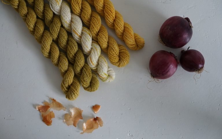 five mini skeins of yarn naturally dyed with onion skins alongside some yellow onion skins and three red onions